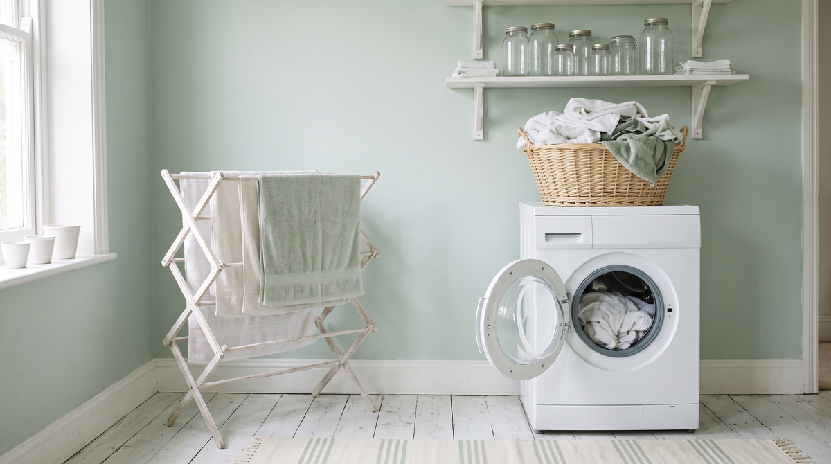 A pale mint laundry room with a white washing machine, drying rack of towels and a wicker basket of linens