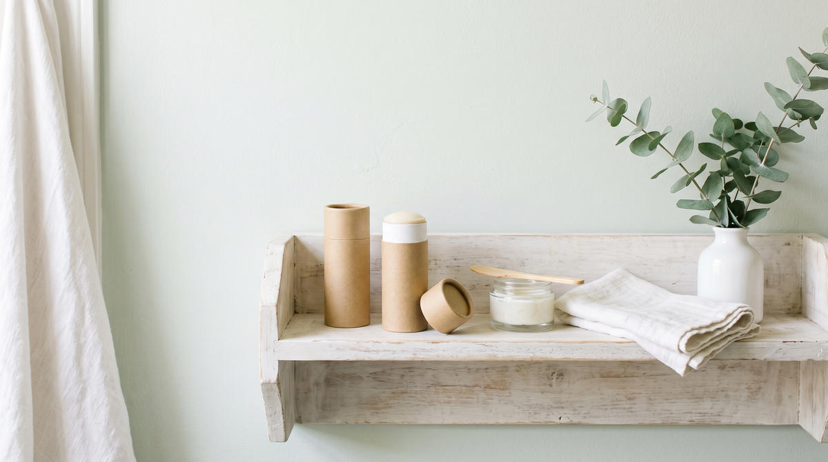 A whitewashed shelf with blank kraft-paper deodorant tubes, one open showing cream, a glass jar and eucalyptus in a white vase