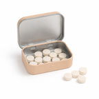 An open beige tin filled with off-white round tablets, with four additional tablets sitting on the white surface nearby.