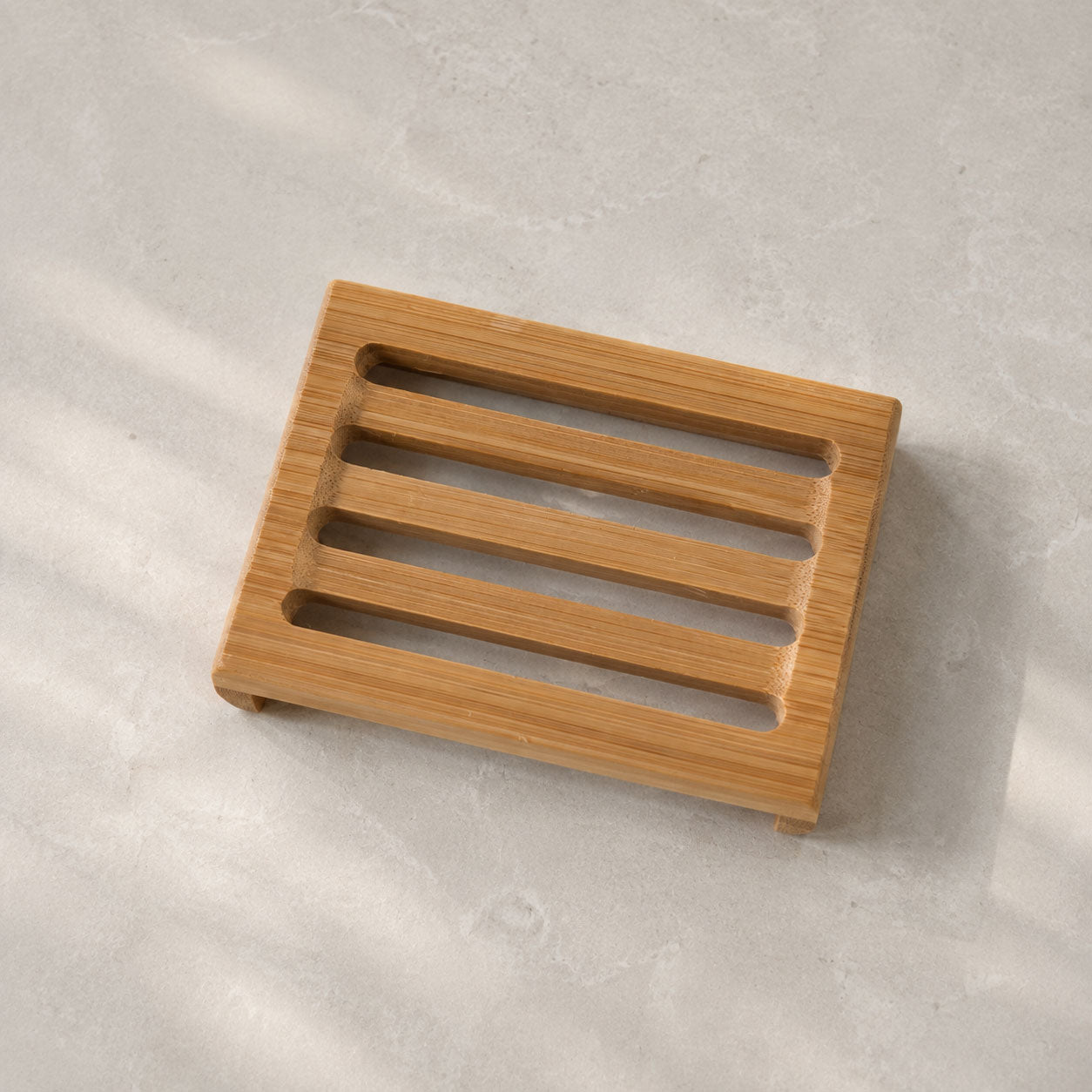 Slotted rectangular bamboo soap tray resting on a light grey marble countertop in natural sunlight.