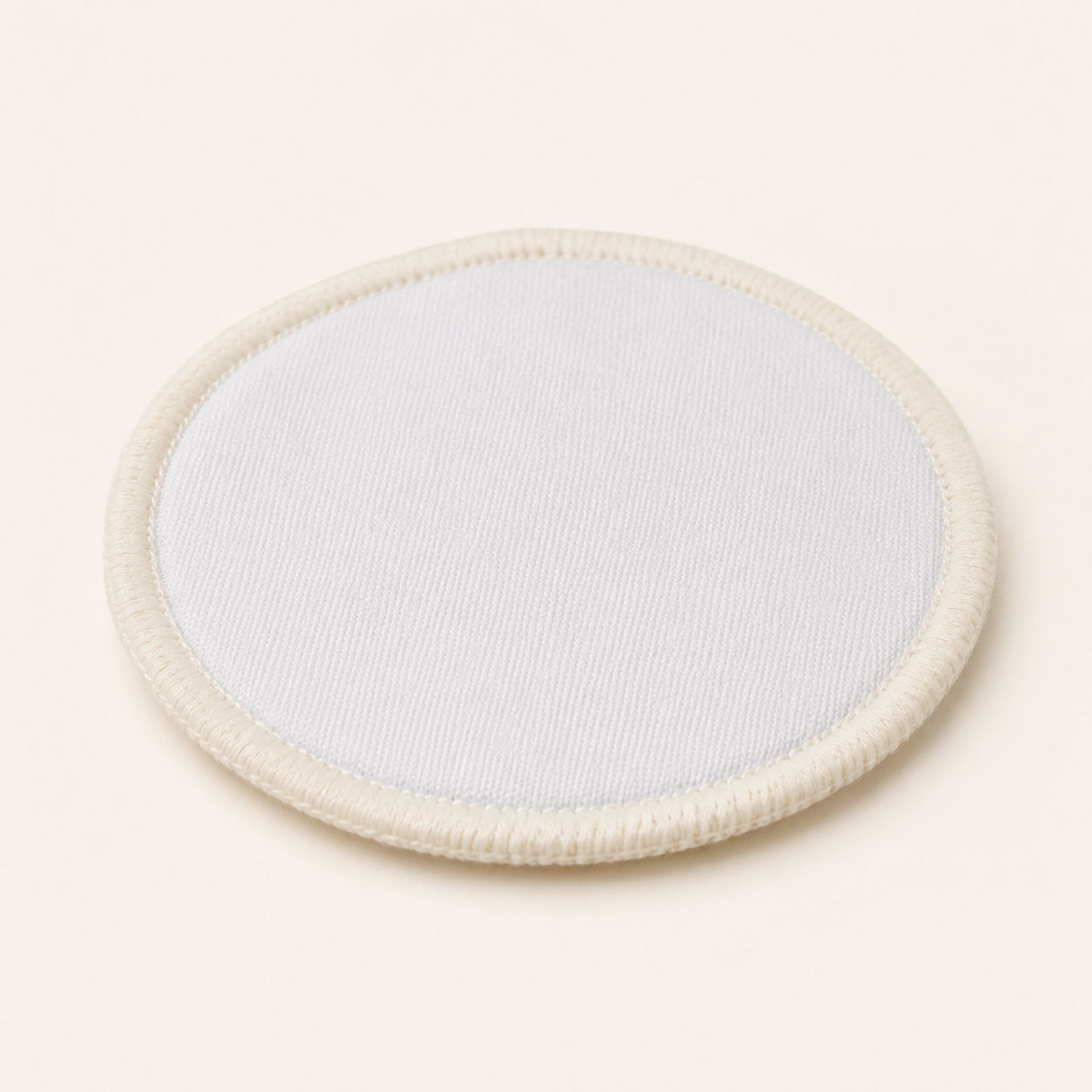 Single circular white reusable cotton makeup pad with stitched edge on an off-white background.
