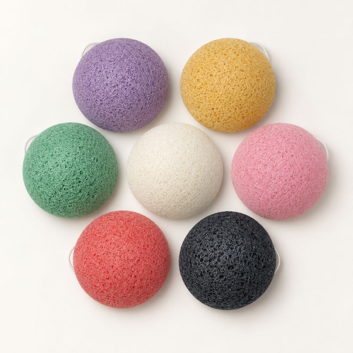 Seven colourful, dome-shaped konjac sponges arranged in a circle on a white surface, viewed from above.
