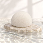 White konjac sponge resting on a flat textured stone in shallow, clear rippling water.