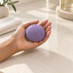 A person holds a purple textured konjac facial sponge in an open hand over a bathroom counter.