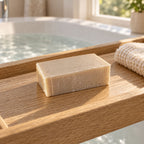 Rectangular beige soap bar resting on a wooden bath tray next to a rolled cloth over a filled tub.
