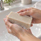 Hands lathering the rectangular beige soap bar with white foam over a sink.
