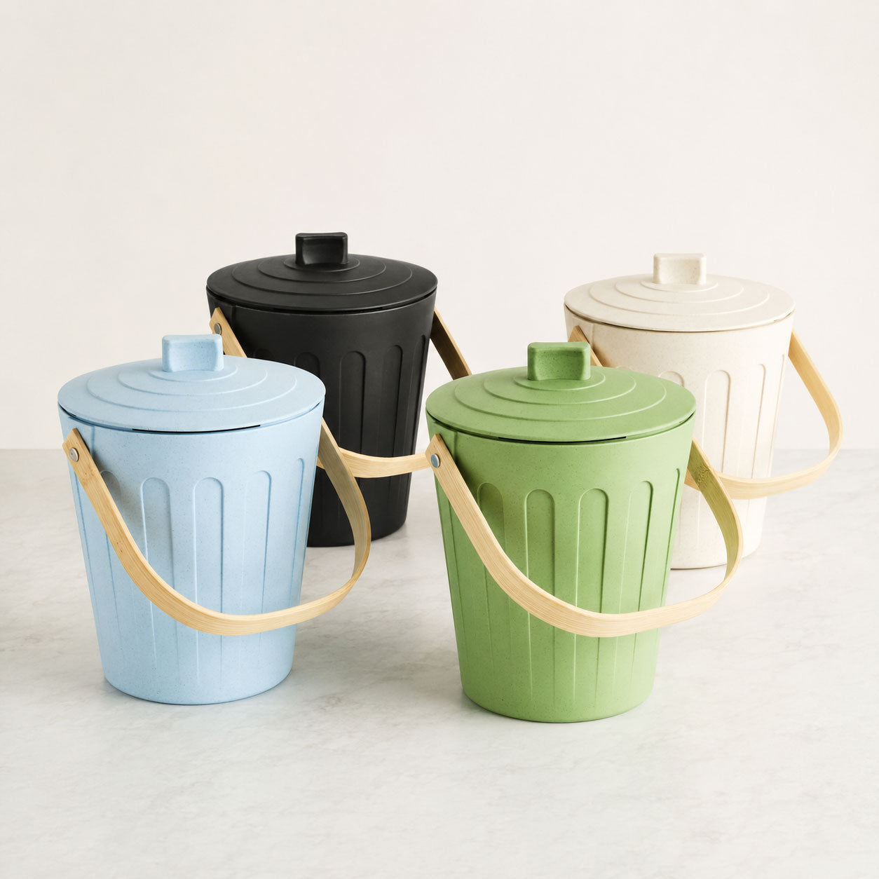 Four Bamboozle by Martha Stewart composter bins with ribbed bodies, screw-top lids, and light bamboo handles, shown in light blue, green, black, and off-white on a light surface against a pale background.