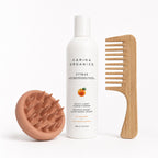 Carina Organics Citrus Daily Light Conditioner bottle standing between a clay pink silicone scalp massager and a bamboo wide-tooth detangling comb on a white background