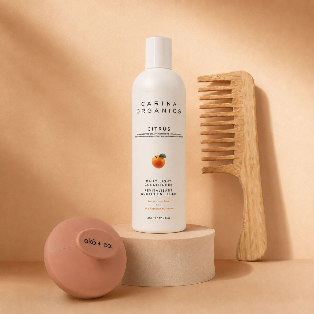 Carina Organics citrus conditioner raised on a stone plinth with a bamboo wide-tooth comb leaning behind it and an ekö + co. branded clay pink scalp massager resting in front