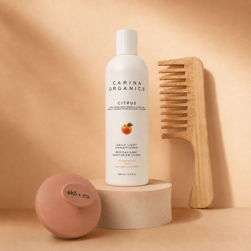 Carina Organics citrus conditioner raised on a stone plinth with a bamboo wide-tooth comb leaning behind it and an ekö + co. branded clay pink scalp massager resting in front