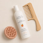 Flat lay of the Carina Organics Citrus Daily Light Conditioner with a clay pink silicone scalp massager and a long-handled bamboo wide-tooth comb on a cream background