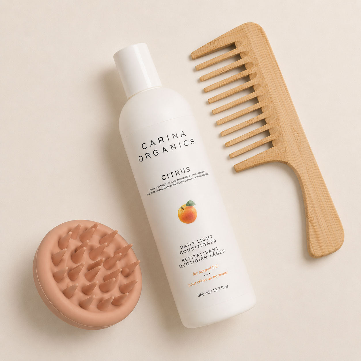 Flat lay of the Carina Organics Citrus Daily Light Conditioner with a clay pink silicone scalp massager and a long-handled bamboo wide-tooth comb on a cream background