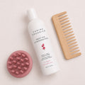 Flat lay of the Carina Organics Sweet Pea Daily Light Conditioner with a pink silicone scalp massager and a handle-free wide tooth bamboo comb on a cream background
