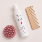 Flat lay of the Carina Organics Sweet Pea Daily Light Conditioner with a pink silicone scalp massager and a handle-free wide tooth bamboo comb on a cream background
