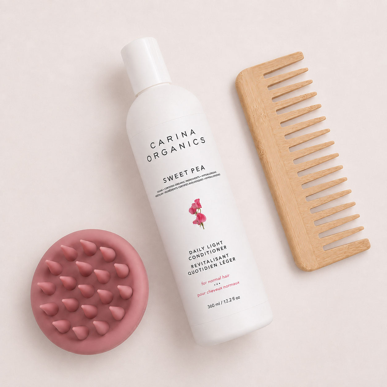 Flat lay of the Carina Organics Sweet Pea Daily Light Conditioner with a pink silicone scalp massager and a handle-free wide tooth bamboo comb on a cream background