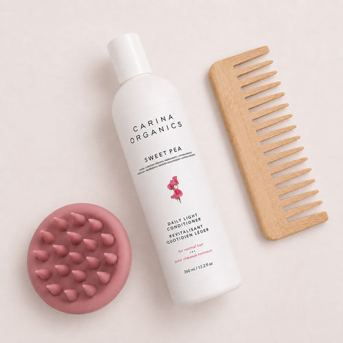 Flat lay of the Carina Organics Sweet Pea Daily Light Conditioner with a pink silicone scalp massager and a handle-free wide tooth bamboo comb on a cream background