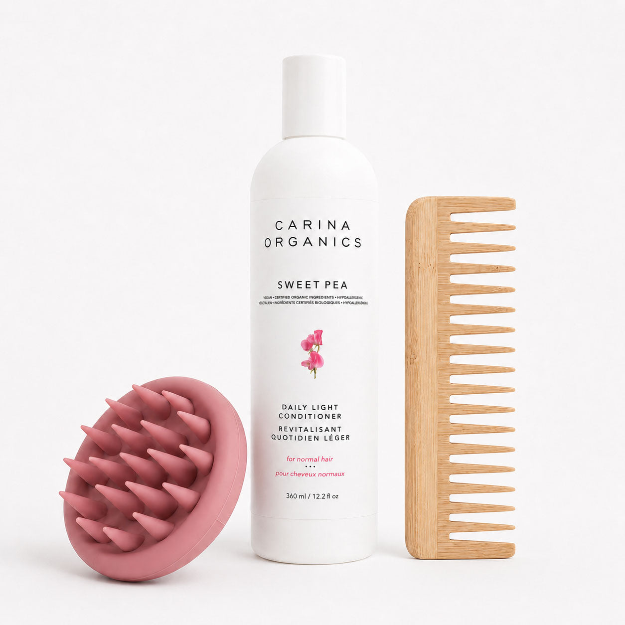 Carina Organics Sweet Pea Daily Light Conditioner bottle standing between a pink silicone scalp massager and a rectangular wide tooth bamboo comb on a white background