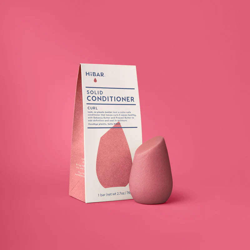 A pink, teardrop-shaped HiBAR Curl solid conditioner bar stands next to its white and pink paper carton on a pink background.