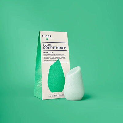 A light green, teardrop-shaped solid conditioner bar stands next to its triangular HiBAR packaging on a green background.
