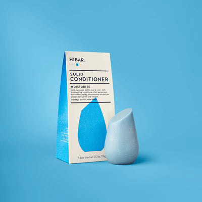 A light blue HiBAR solid conditioner bar stands next to its white and blue triangular paper carton on a blue background.