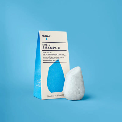 A light-grey, stone-shaped shampoo bar stands next to its blue and white HiBAR carton on a solid blue background.