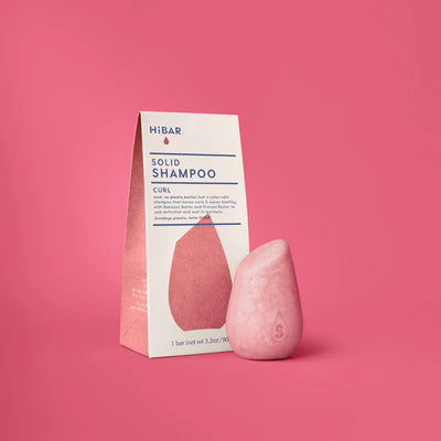 A pink, stone-shaped shampoo bar with an 'S' imprint next to its white HiBAR packaging on a solid pink background.