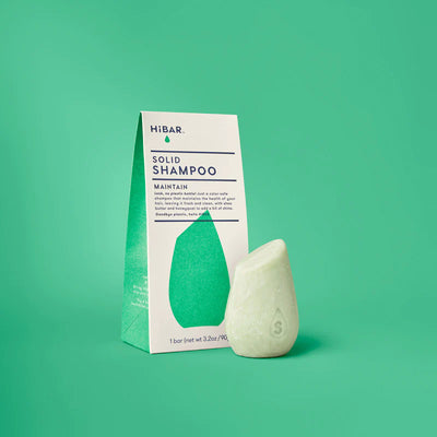 A light green, teardrop-shaped shampoo bar stands next to its white and green HiBAR cardboard packaging.