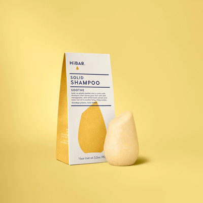A light yellow, teardrop-shaped solid shampoo bar stands beside its triangular white and yellow HiBAR cardboard box.