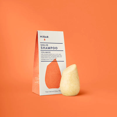 A cream-coloured, teardrop-shaped HiBAR solid shampoo bar next to its orange and white box on an orange background.