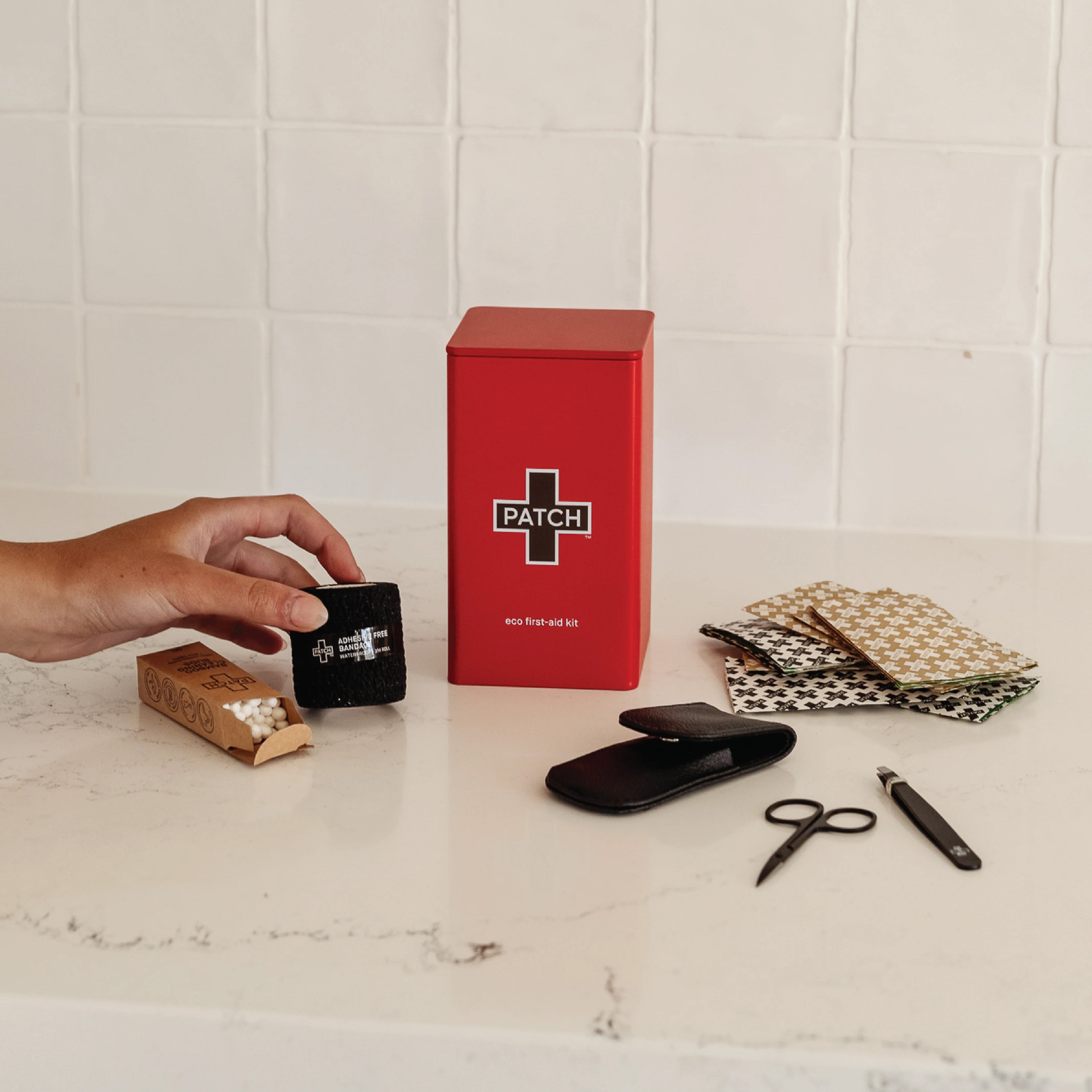 Red PATCH first-aid tin with contents displayed including bamboo bandages with panda prints, black scissors, tweezers, bamboo container, and safety pouch on marble countertop