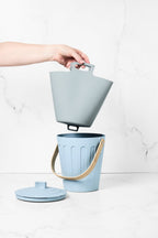 Hand holding a grey Bamboozle Martha Stewart silicone liner bag above a matching light blue compost pail with lid.