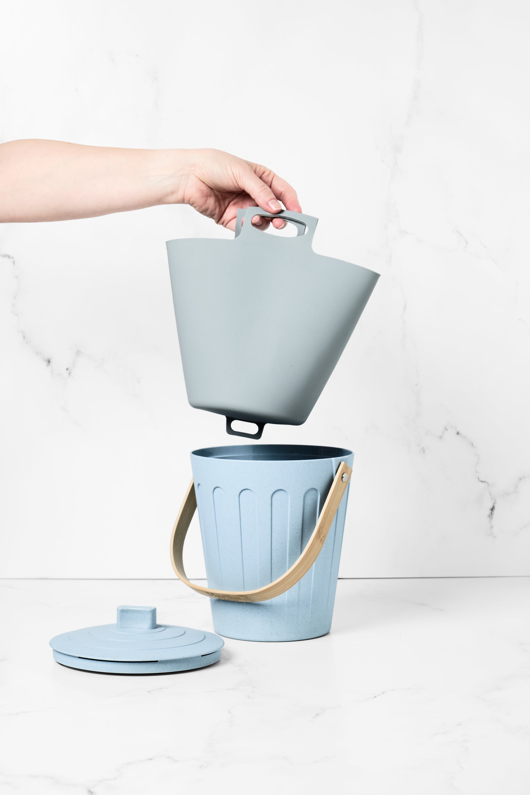 Hand holding a grey Bamboozle Martha Stewart silicone liner bag above a matching light blue compost pail with lid.