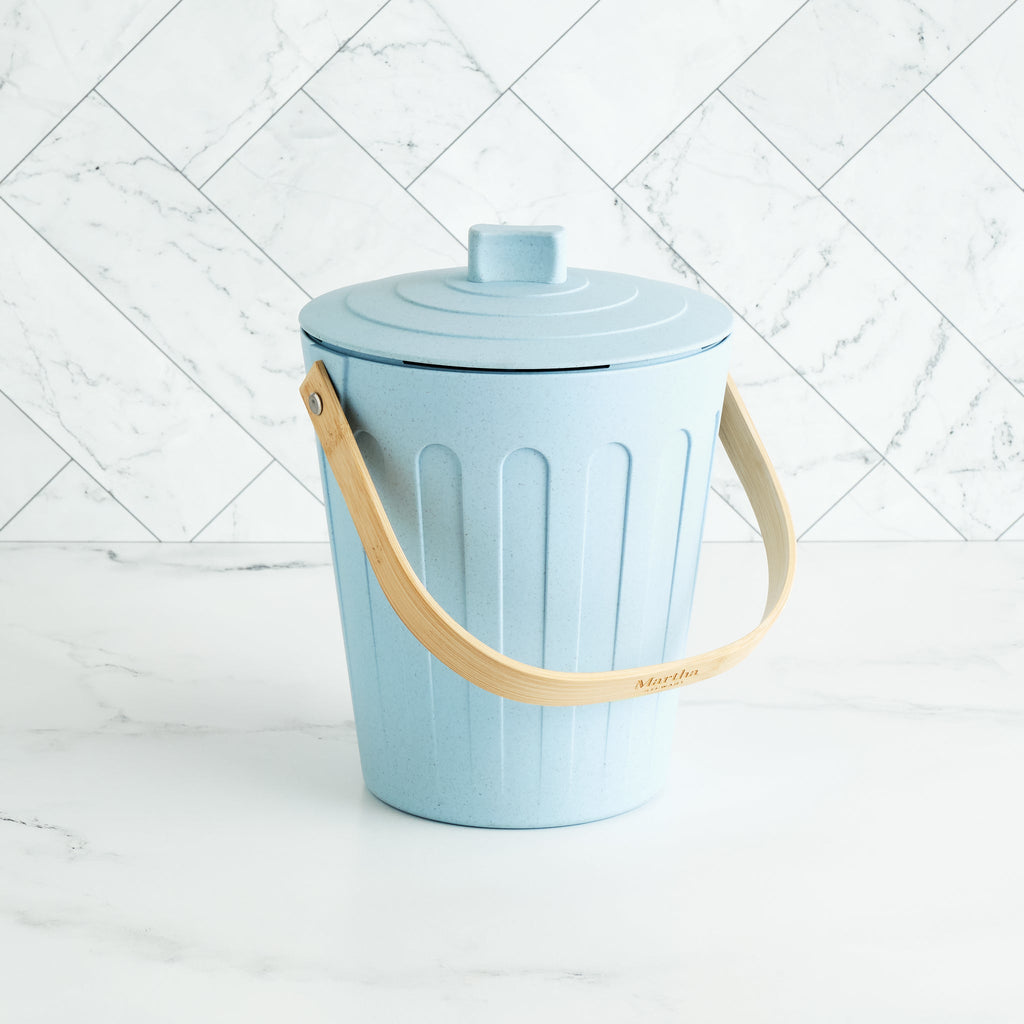Martha Stewart light blue indoor compost bin with a wooden handle on a white marble surface and tile backsplash.
