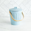 Martha Stewart light blue indoor compost bin with a wooden handle on a white marble surface and tile backsplash.