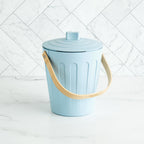 Martha Stewart light blue indoor compost bin with a wooden handle on a white marble surface and tile backsplash.