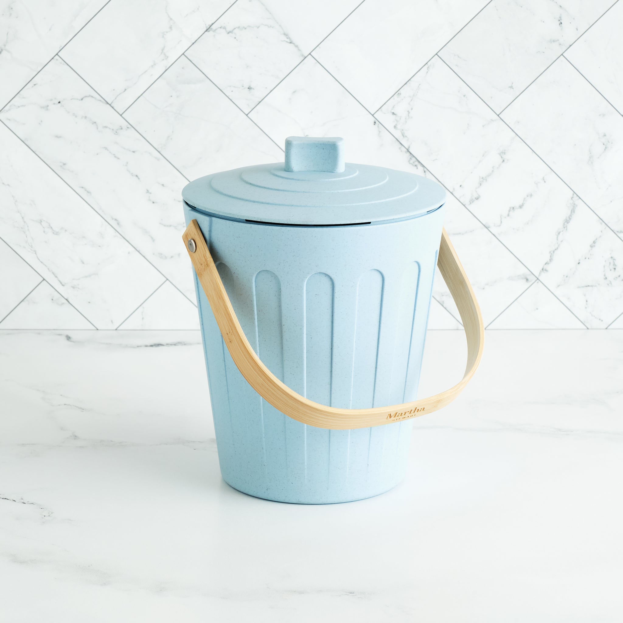 Martha Stewart light blue indoor compost bin with a wooden handle on a white marble surface and tile backsplash.