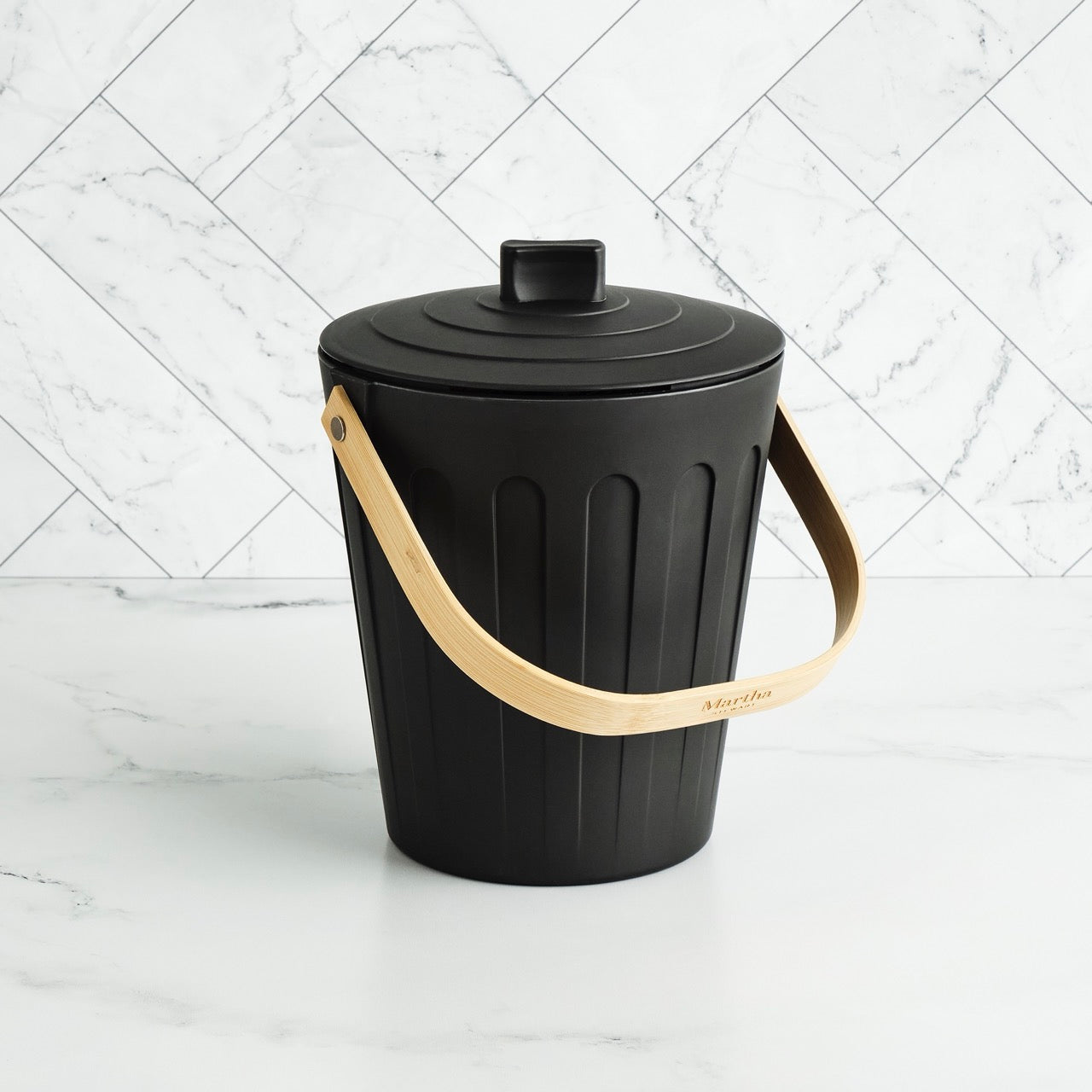 Martha Stewart black indoor compost bin with a wooden handle on a white marble surface and tile backsplash.