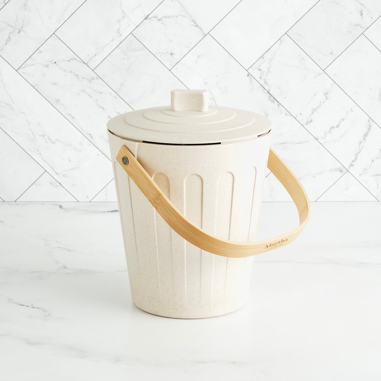 Martha Stewart beige indoor compost bin with a wooden handle on a white marble surface and tile backsplash.