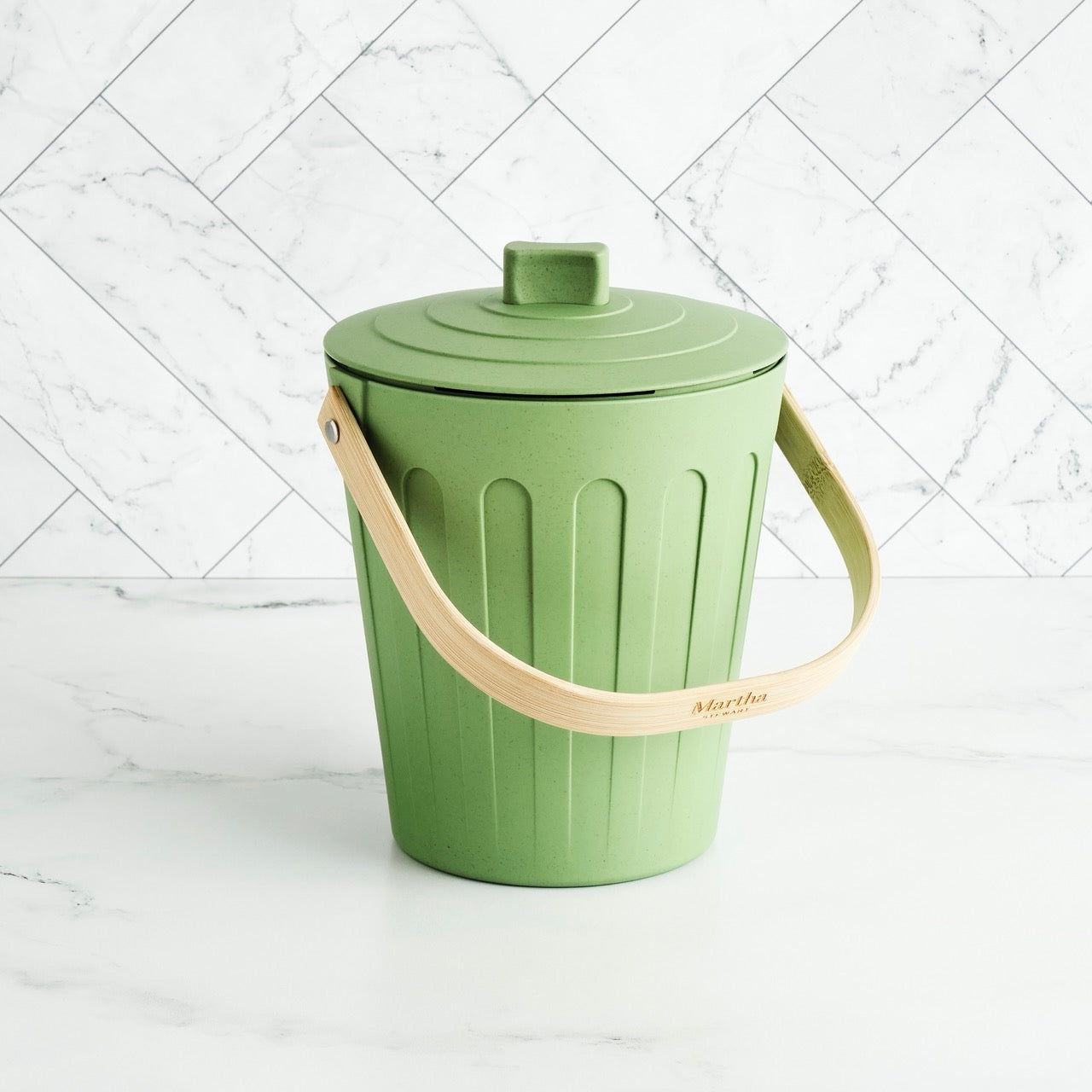 Martha Stewart green indoor compost bin with a wooden handle on a white marble surface and tile backsplash.