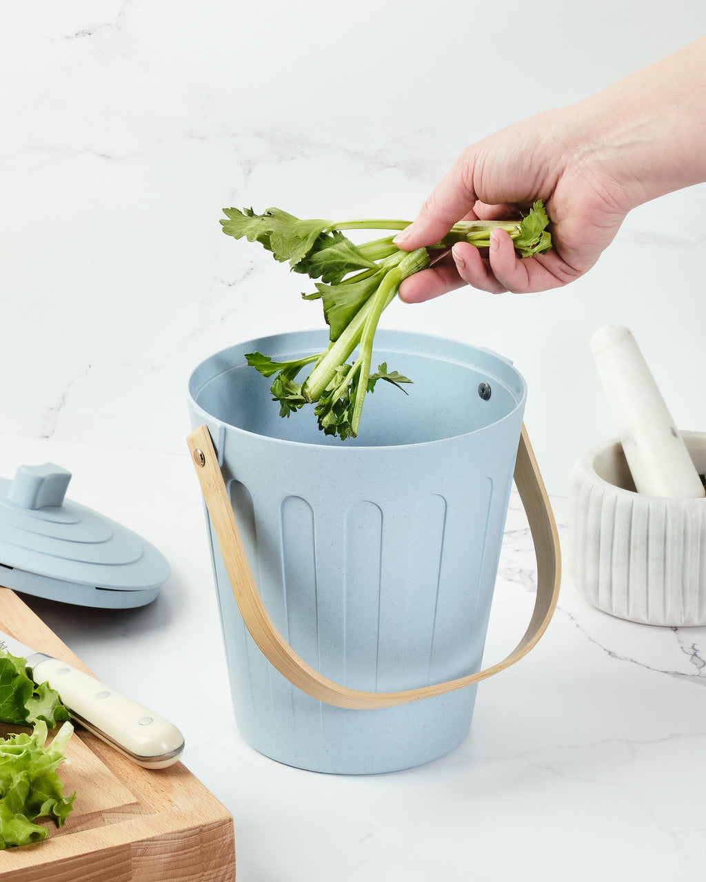 Hand placing celery into a Martha Stewart light blue indoor compost bin on a kitchen counter.