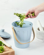 Hand placing celery into a Martha Stewart light blue indoor compost bin on a kitchen counter.