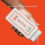 A hand holding a white Native body wash bottle, dispensing clear liquid against an orange backdrop.