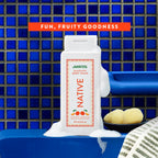 White bottle of Native Mandarin body wash covered in lather on a blue tile shelf beside a bath sponge.