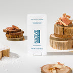 Native deodorant stick surrounded by wooden log slices, cedar shavings, and coarse sea salt crystals.