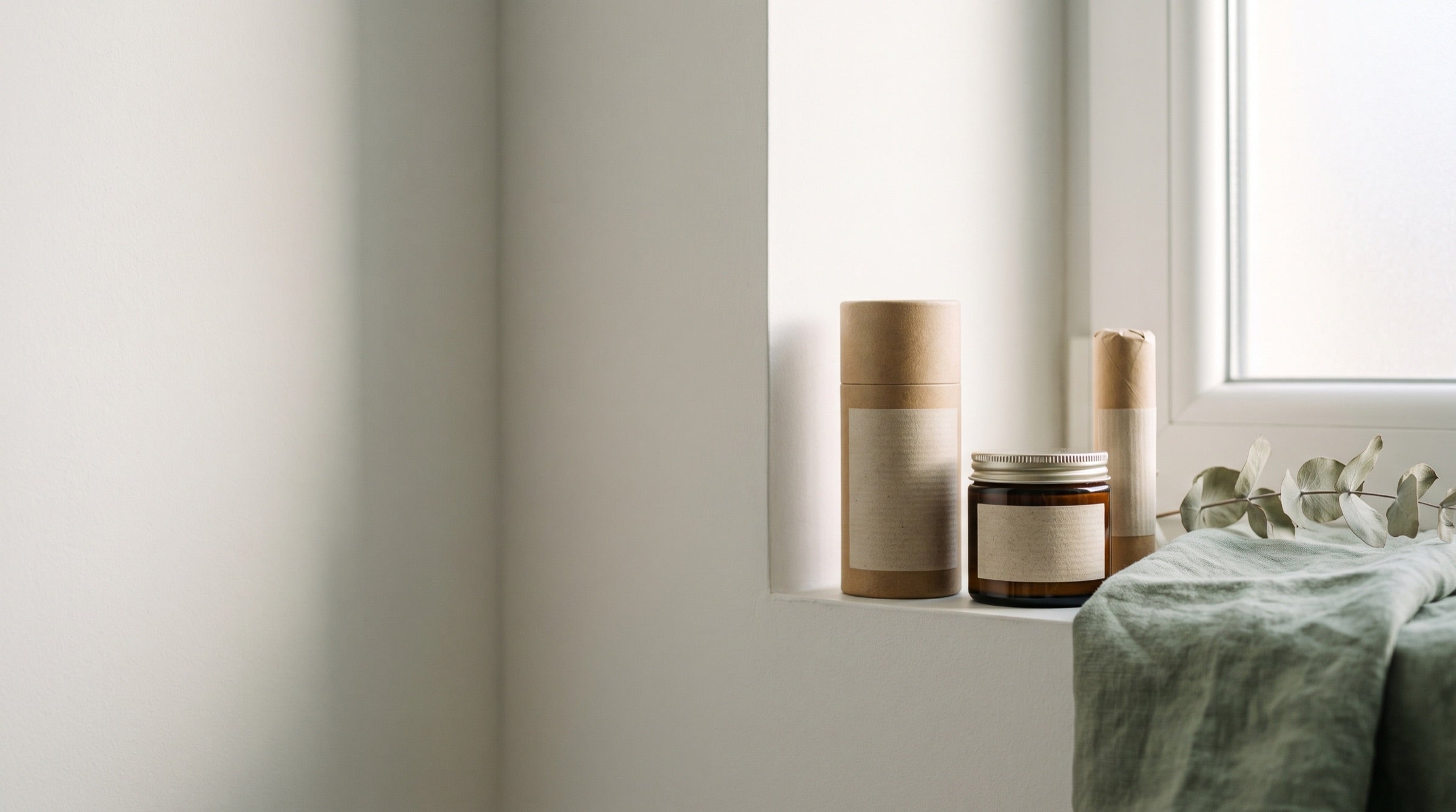 Three plastic-free deodorants — a cardboard tube, an amber glass jar and a paper-wrapped stick — on a sunlit bathroom windowsill