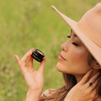 Woman smelling Routine The Class solid perfume - model in hat holding black jar in natural outdoor setting - demonstrating mindful wellness fragrance ritual