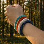 Four coiled repellent bands in teal, orange, white, and navy blue worn stacked on a raised wrist in a sunny forest.