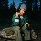 Someone in a green jacket holds a mug on a rock in the forest at dusk, wearing stacked coiled bands on their wrist.