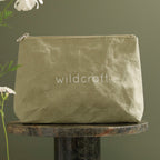 A crinkled olive green Wildcraft pouch sits on a round marble pedestal against a green background with white flowers.