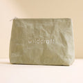 A crinkled olive green Wildcraft pouch stands on a plain beige surface against a warm cream background.